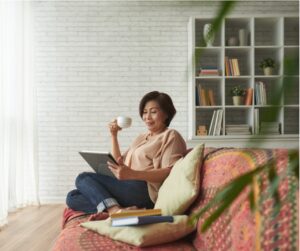 A mature woman of East Asian heritage relaxes on a couch while sipping tea and looking at her laptop
