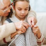 Senior woman teaches little girl how to knit wool threads with spokes. Closeup image